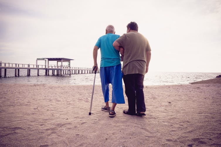Een jongere man ondersteunt een oudere man die op het strand loopt.