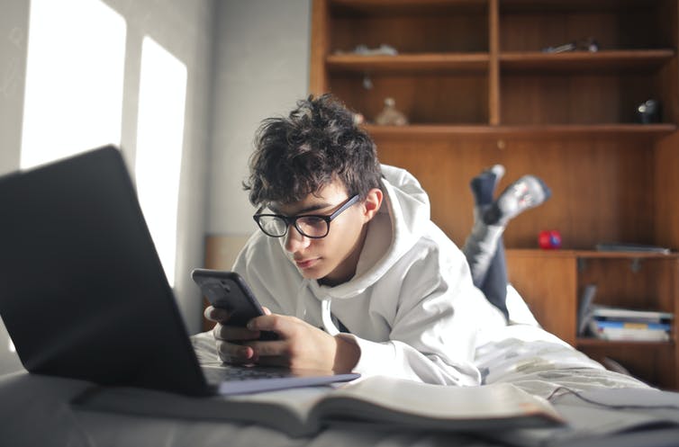 Een tienerjongen ligt op zijn bed met zijn smartphone, met een open laptop.