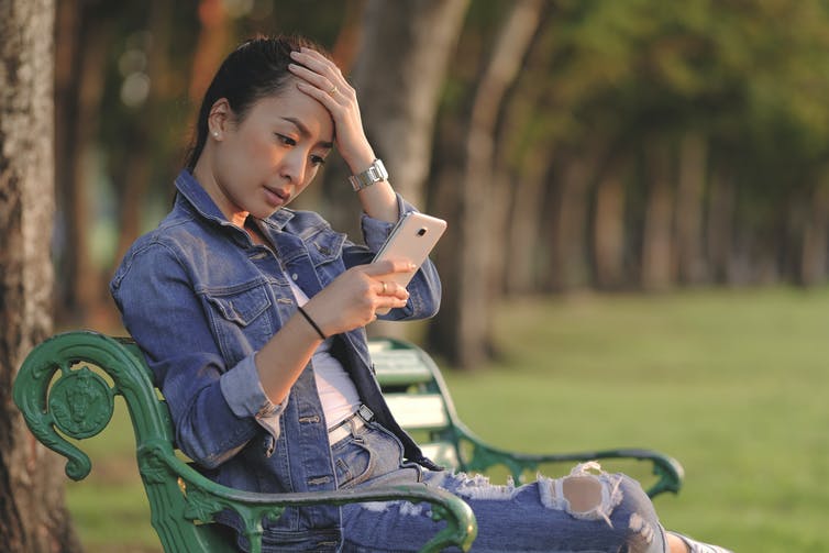 Een jonge vrouw zit op een bankje in het park en kijkt naar haar smartphone.  Ze lijkt gespannen.