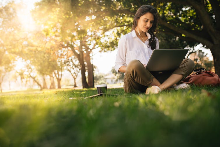 Een vrouw zit in een park naar iets te kijken op haar laptop, met oordopjes in.