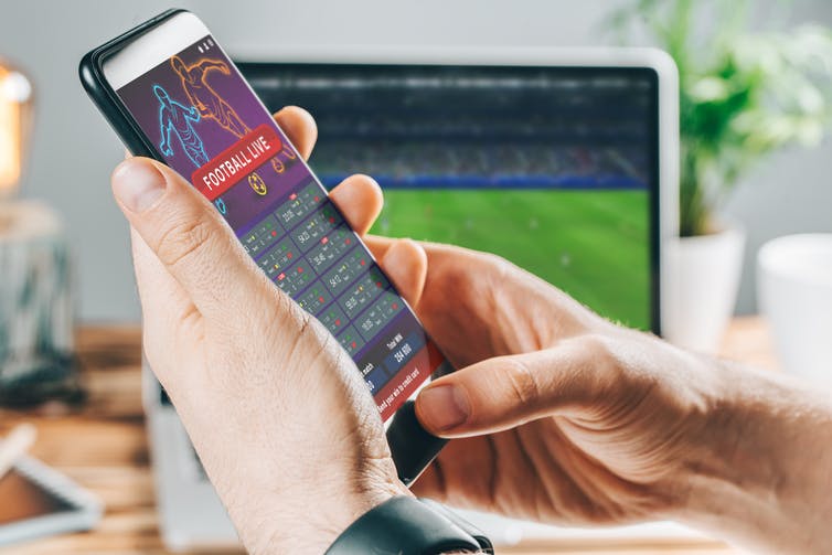 Een hand houdt een smartphone vast met een app voor sportweddenschappen open, voor een laptopscherm waarop een sportwedstrijd wordt getoond.
