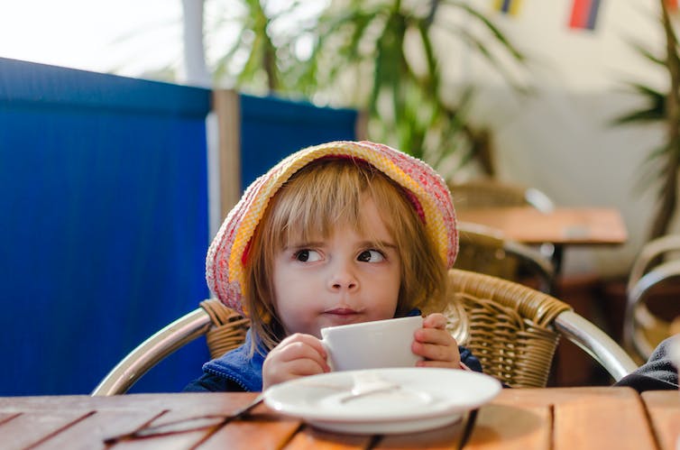 Klein meisje met een slappe hoed tuurt over een tafel, houdt een koffiemok vast met een brutale uitdrukking