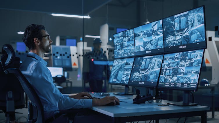 Een man zit aan een bureau voor een reeks schermen die elk beelden van CCTV-camera's tonen.