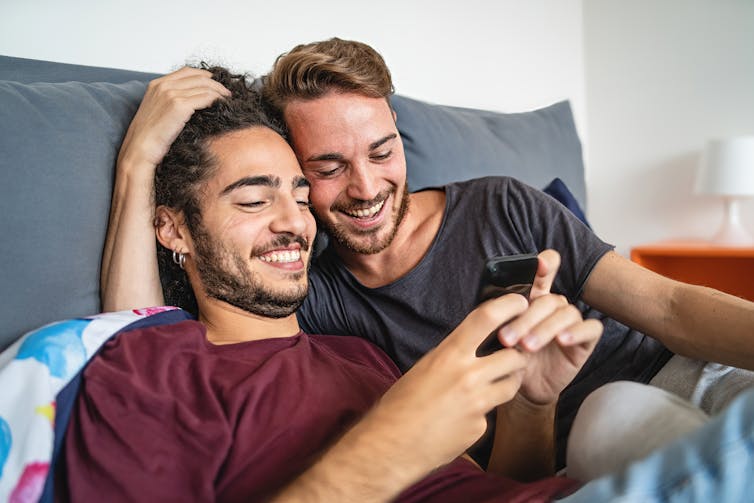 Een homostel samen in bed, blij kijkend naar een telefoon