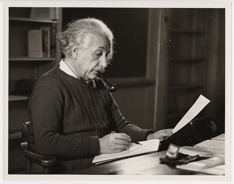 Albert Einstein zittend aan zijn bureau met een pijp die papieren markeert.